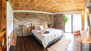 A bedroom with a wooden ceiling, a brick wall, a bed with pillows, a nightstand, and a chair.
