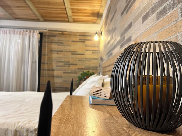 A modern bedroom with a wooden ceiling, a brick wall, a round black vase on a wooden table, and a bed with white bedding. - Autenthico Sajonia Hotel Aeropuerto (Rionegro)
