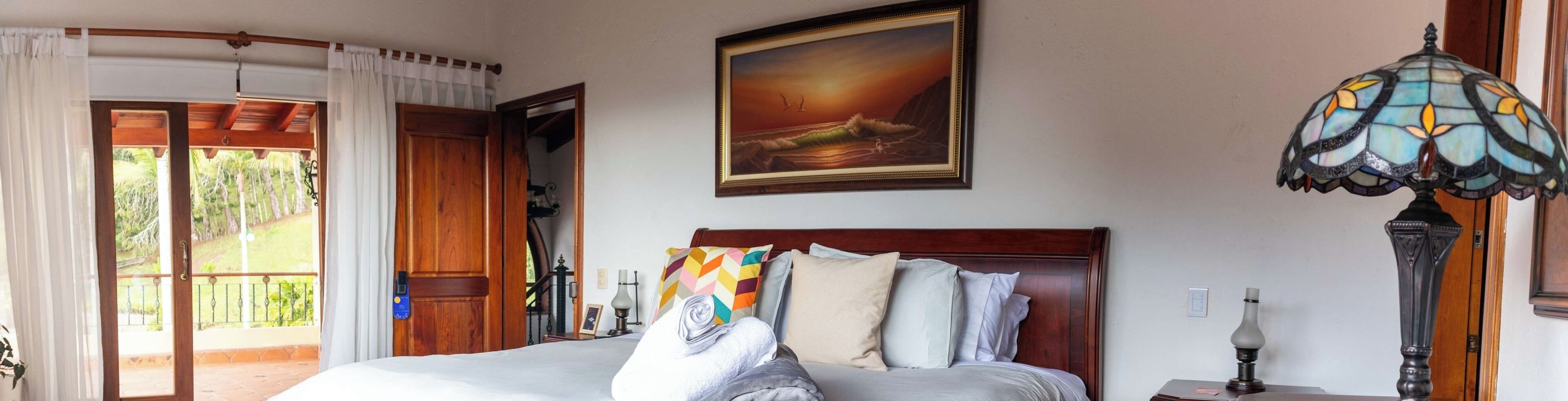 A hotel room with a bed, a painting, a stained glass lamp, and a view of the outdoors.
