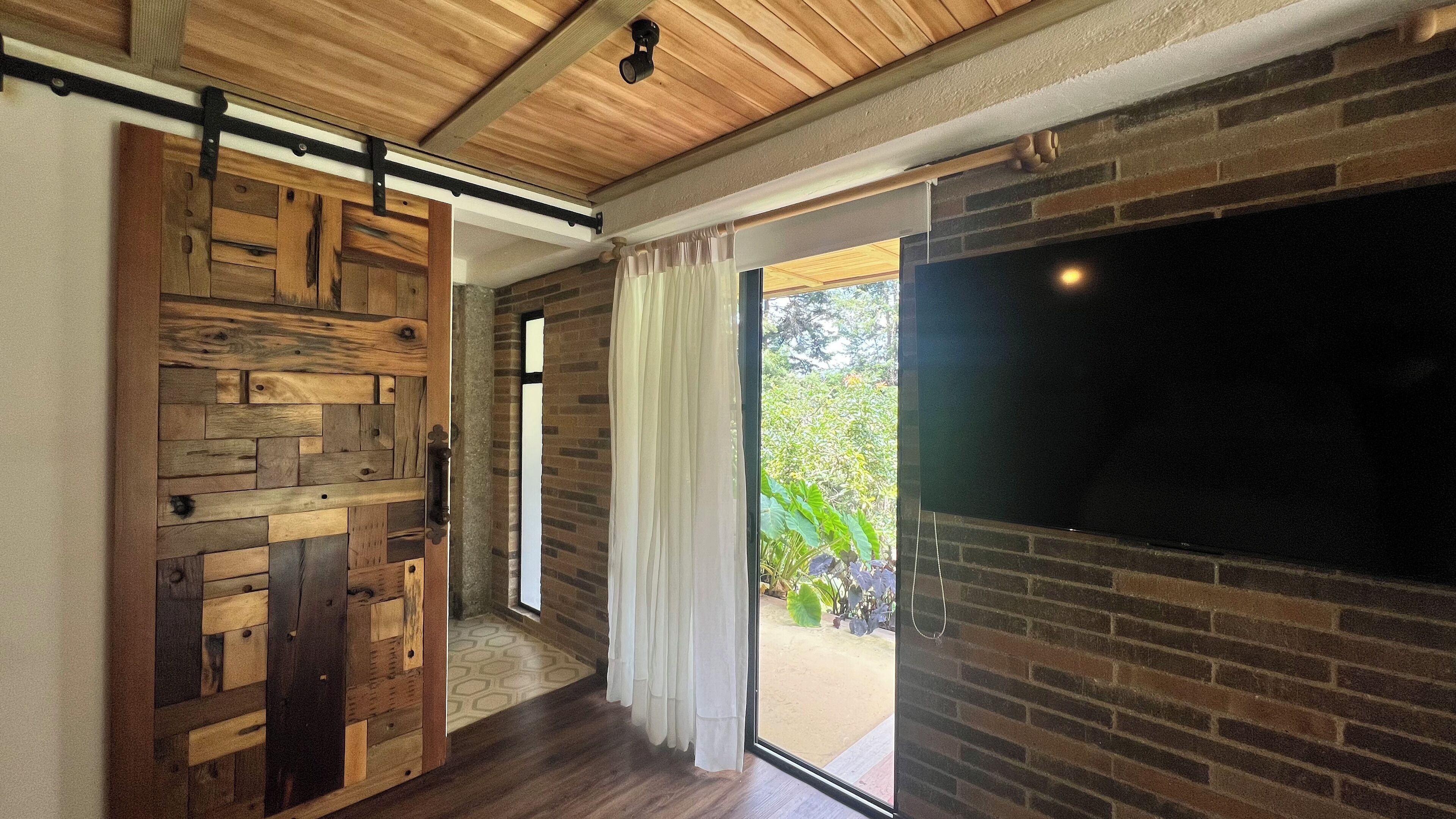 Una habitación con techo de madera, puerta corrediza tipo establo, pared de ladrillo y una gran pantalla de televisión.