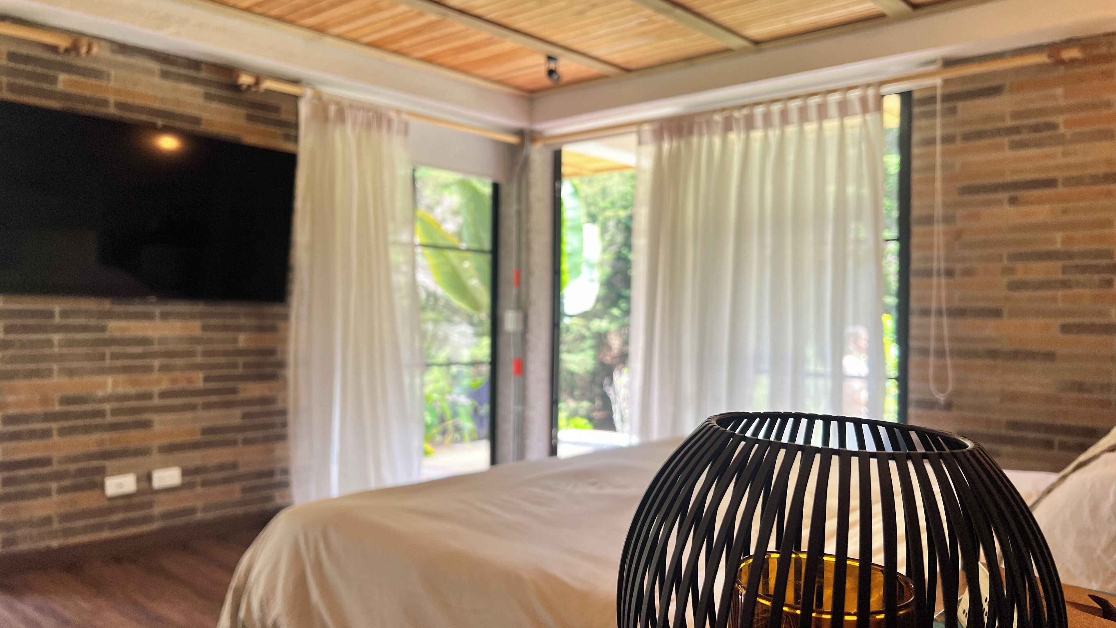 A bedroom with a large bed, a TV mounted on the wall, and a view of greenery through the windows.
