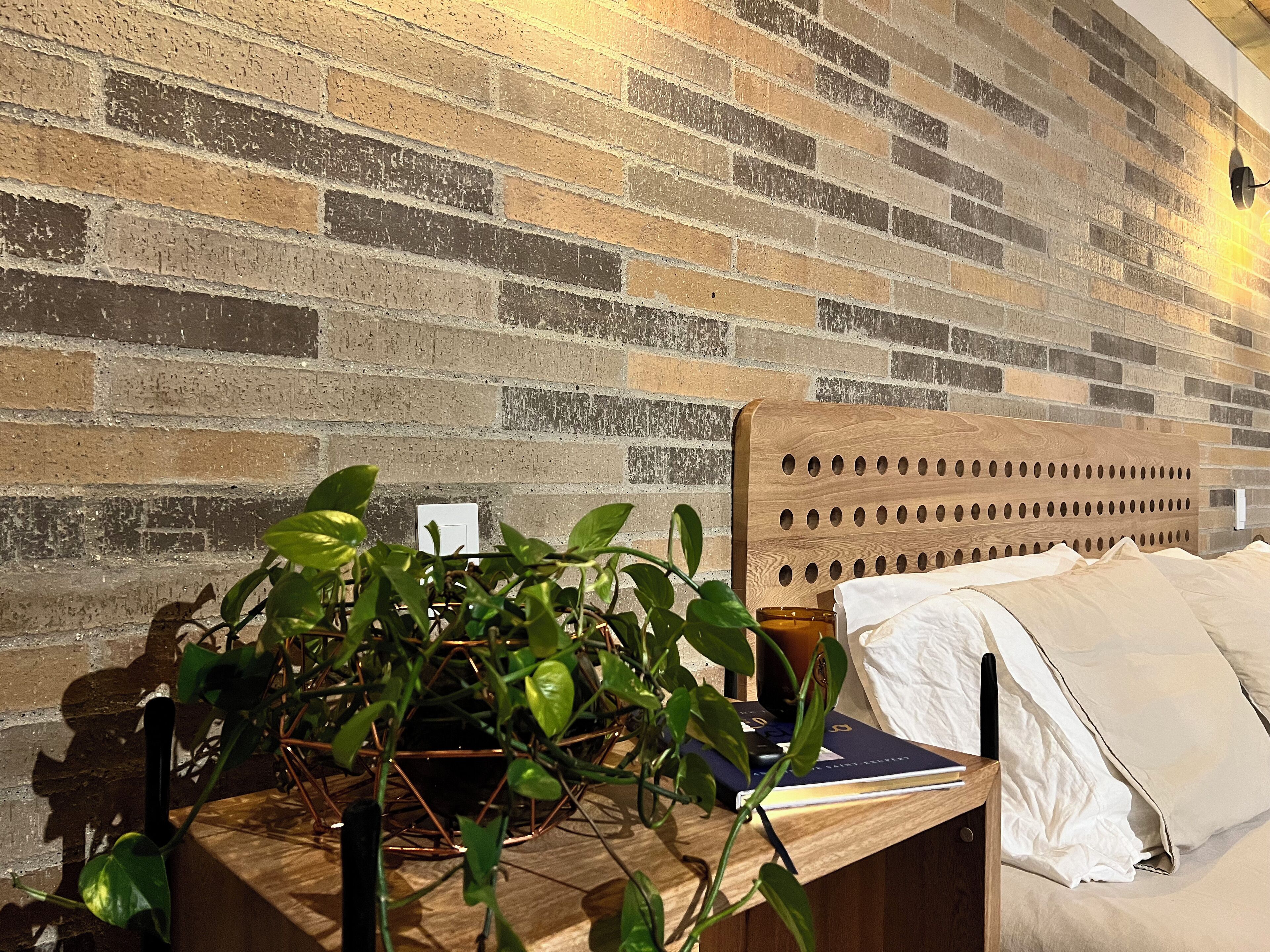 A room with a brick wall, a wooden headboard, a bedside table with a plant, and a bed with white bedding.