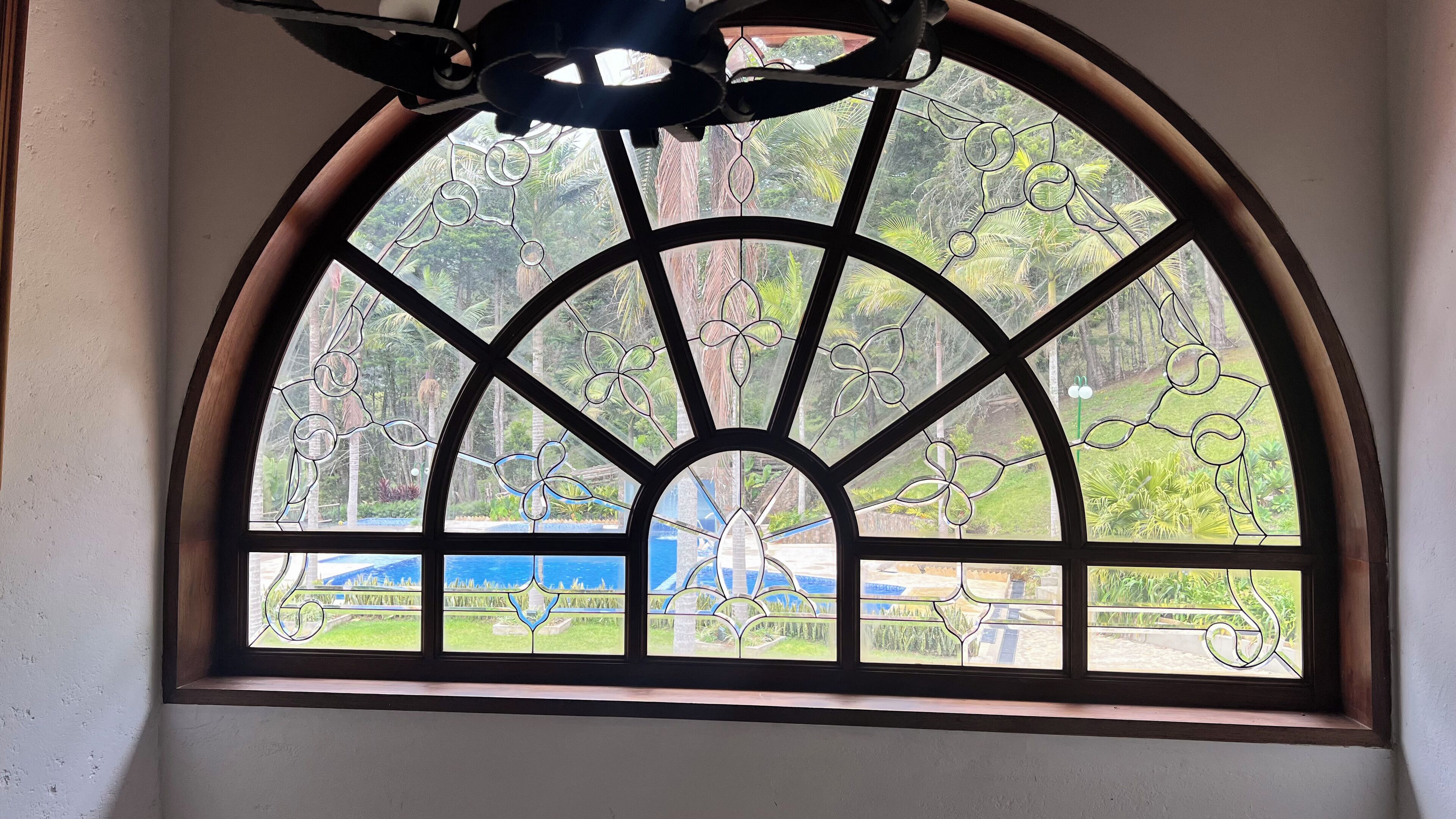 Un amplio ventanal en arco con ornamentación metálica detallada, que ofrece vistas a un jardín exuberante y una piscina.
