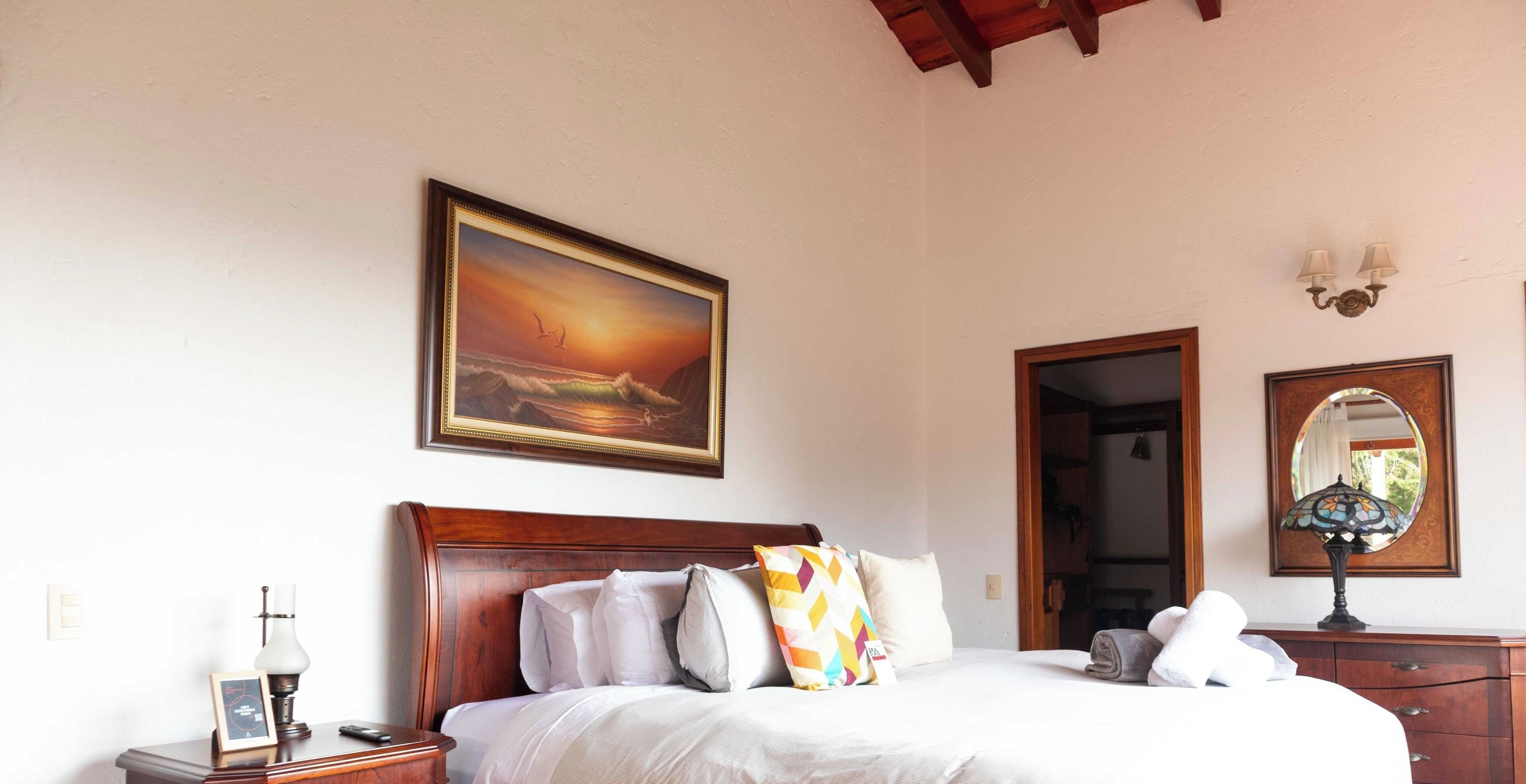 A hotel room with a bed, a nightstand, a lamp, a framed painting, a mirror, and a door leading to another room.