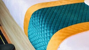 Un letto con una coperta trapuntata color petrolio e un bordo giallo, con cuscini color petrolio e beige.