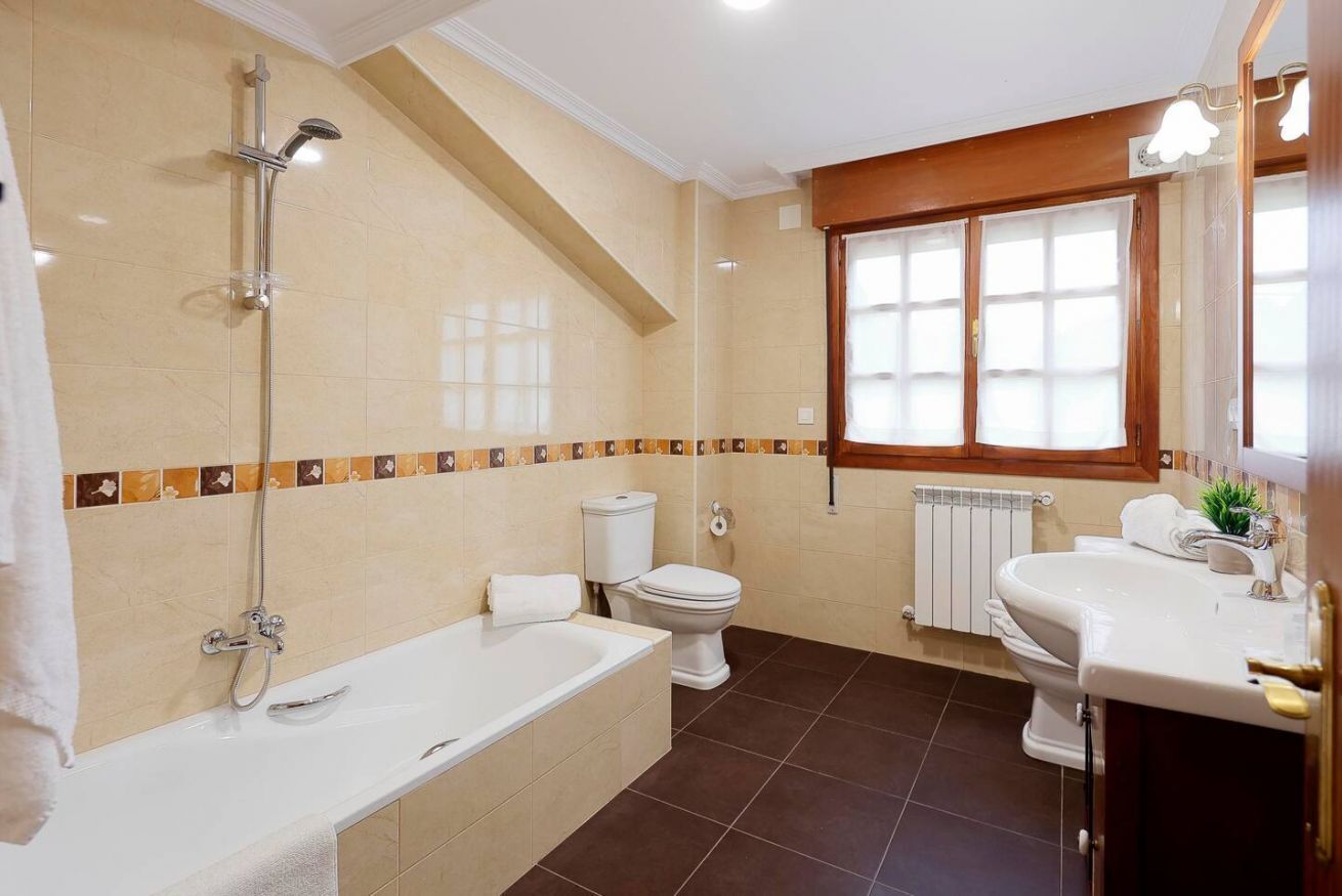 Une salle de bain  équipée d’une baignoire, de toilettes et d’un lavabo, avec un carrelage au sol et une fenêtre avec des stores.