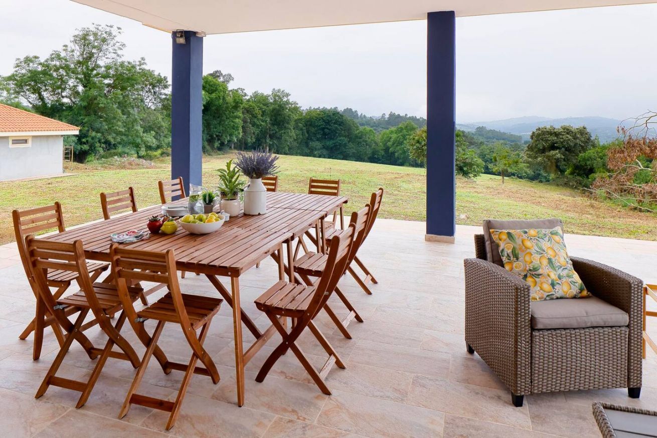 Une terrasse extérieure avec une table et des chaises en bois, un canapé en osier et une vue sur un paysage verdoyant.