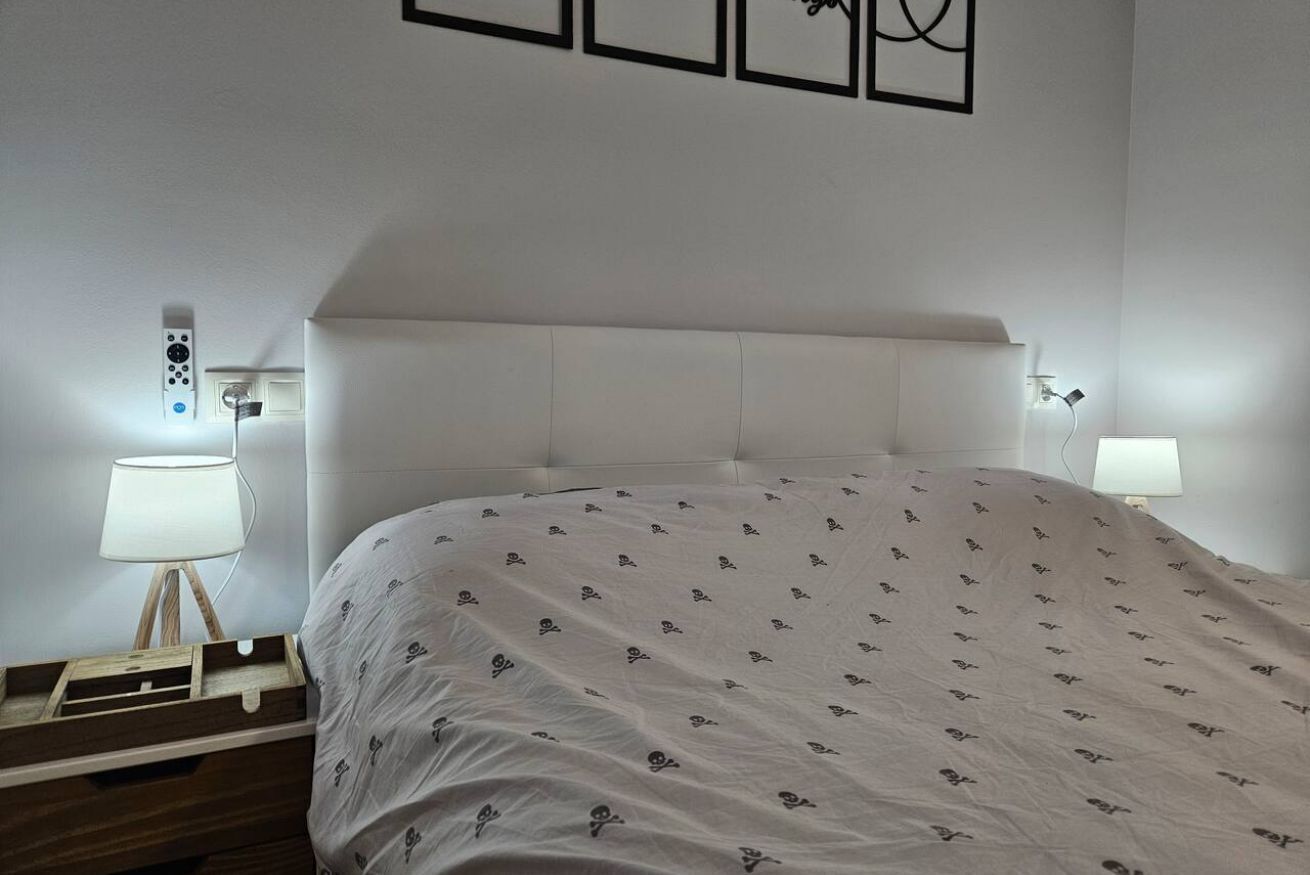 Uma cama bem arrumada com colcha estampada, uma mesinha de cabeceira com abajur e um painel de controle fixado na parede.