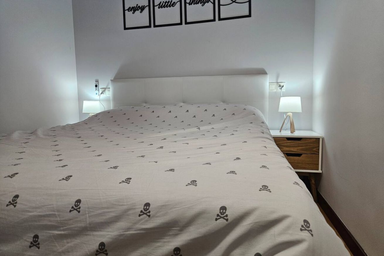 En snyggt bäddad säng med ett mönstrat överkast, ett sängbord med en lampa och en vägg med inramade konstverk.