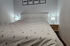 Un lit bien fait, recouvert d’une couvre-lit à motifs, une table de chevet avec une lampe, et un mur orné de tableaux encadrés.