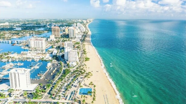 Un paisaje urbano costero con un puerto deportivo, hoteles frente a la playa y un mar azul cristalino.