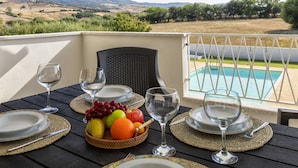 Una mesa de comedor dispuesta con platos, cubiertos y una cesta de frutas, con vista a una piscina.