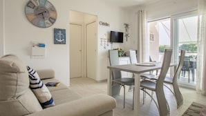 Una sala moderna con un sofá, una zona de comedor con una mesa y sillas, un reloj en la pared y vistas a través de un ventanal.