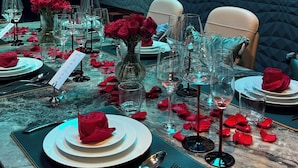 A dining table set with wine glasses, plates, and rose petals, with a cityscape painting in the background. - Space Design Hotel (Dalian)