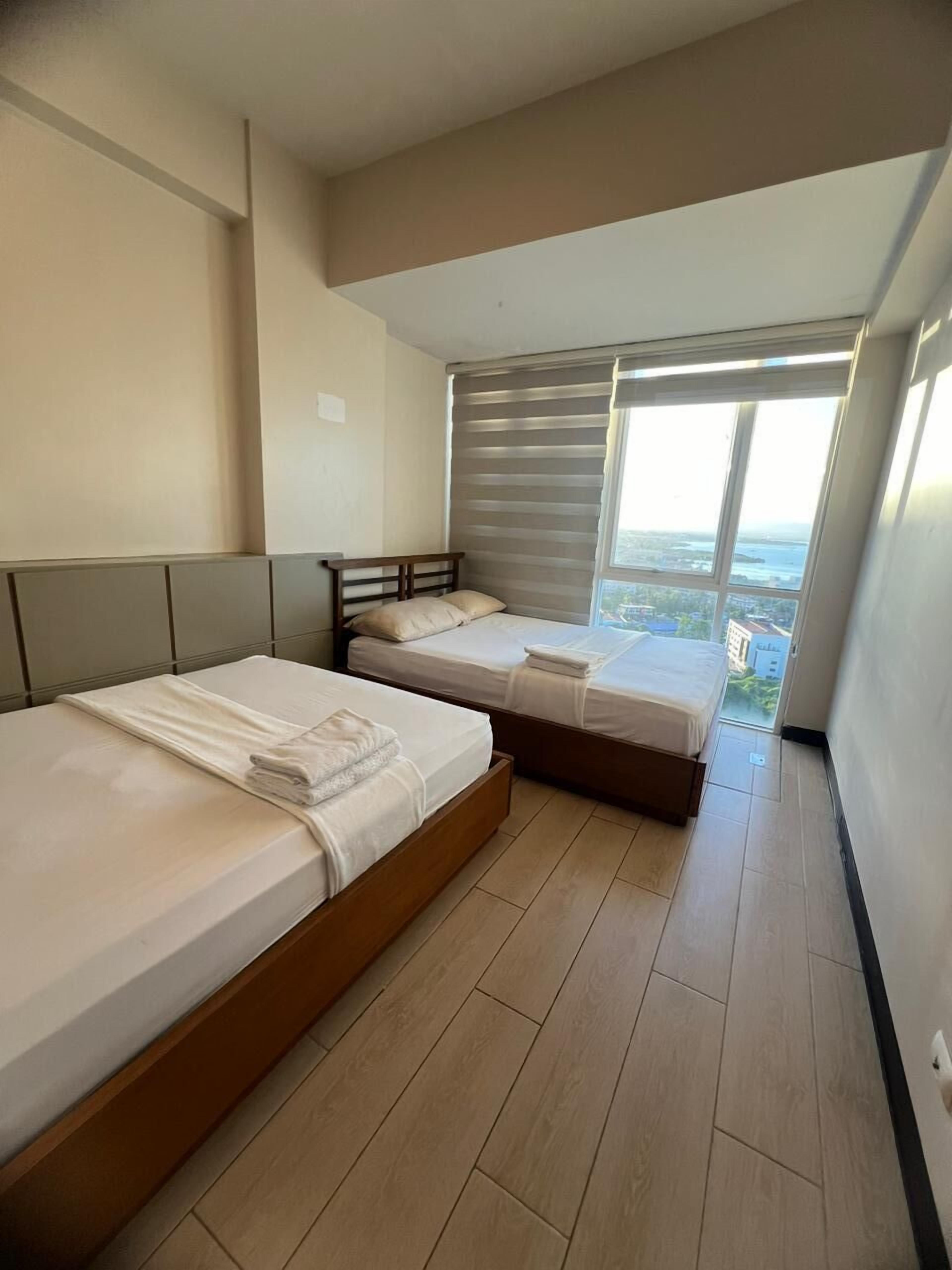 Un dormitorio con dos camas, un ventanal con persianas y vistas a un paisaje urbano.
