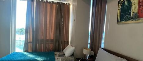A hotel room with a large bed, a smaller bed, a window with curtains, a painting on the wall, and a nightstand with a lamp.