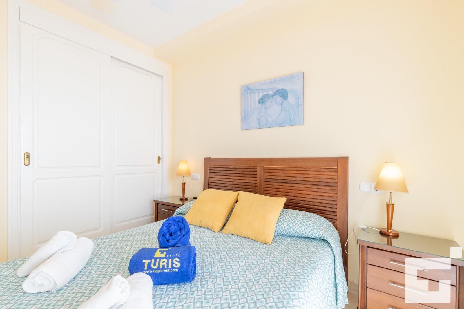 Una cama bien hecha con una colcha estampada, dos almohadas y una toalla azul con la marca "TURIS". Hay dos lámparas de noche, una mesita de noche y un cuadro en la pared.