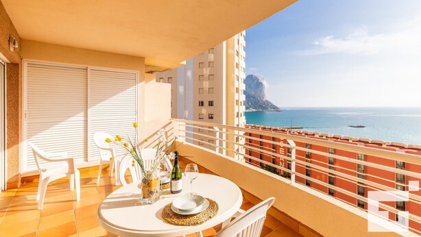 A balcony with a table set for two, overlooking a coastal view with buildings and a prominent rock formation.