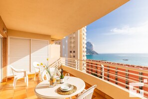 A balcony with a table set for two, overlooking a coastal view with buildings and a prominent rock formation. - Playa de Oro 8B - Grupo Turis - Calpe, Costa Blanca (Calpe)