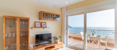 Amplia sala de estar con televisor grande, muebles de madera y balcón con vista al mar.