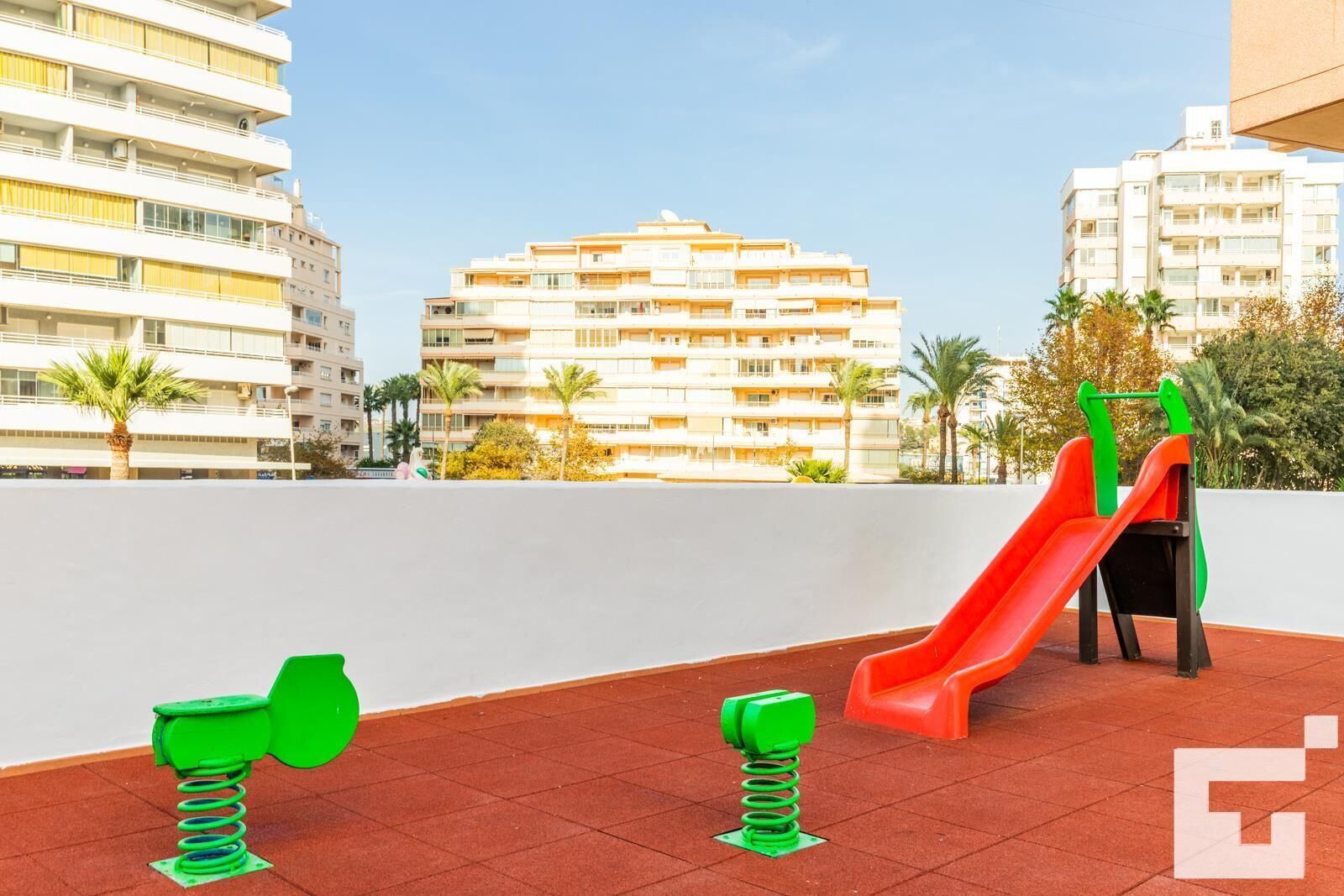 A playground with a slide and swings on a rooftop terrace, surrounded by palm trees and modern buildings.