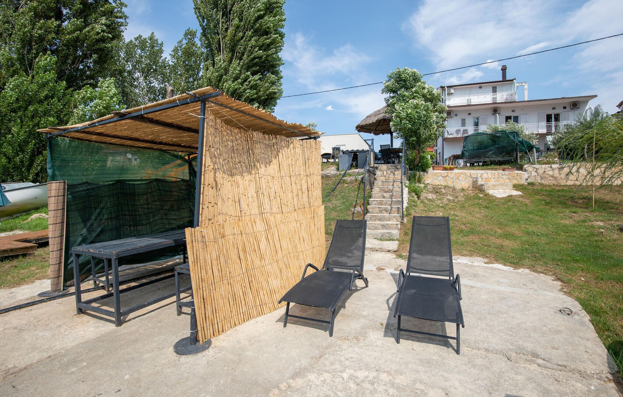 A covered outdoor seating area with a table and two chairs, a thatched roof structure, and a white building in the background.