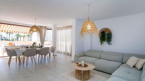 Una sala de estar moderna con un sofá gris, una zona de comedor con mesa y sillas, y dos lámparas colgantes de mimbre.