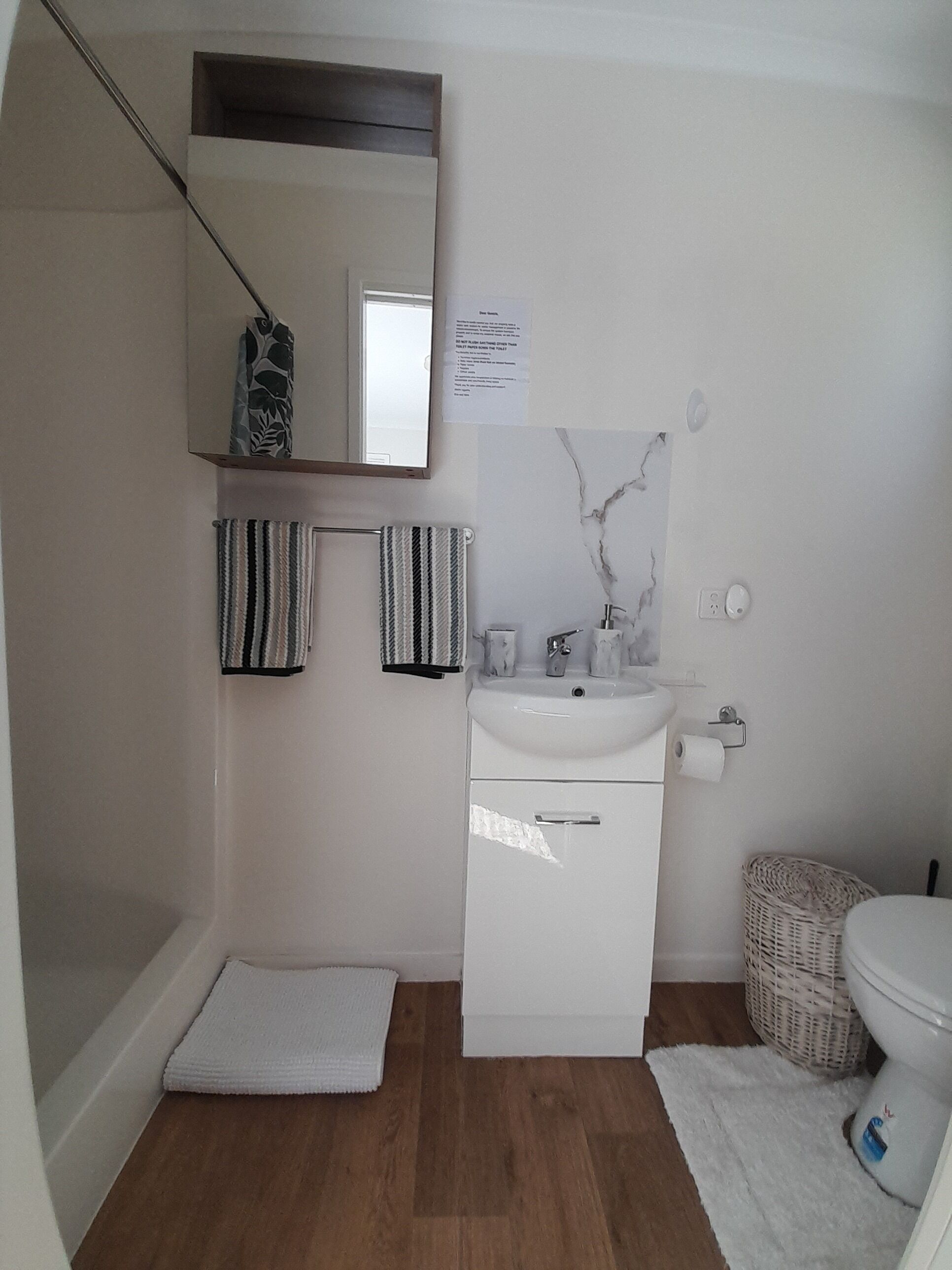 A small, modern bathroom with a white sink, mirror, and toilet. There are two striped towels hanging on the wall, a small wicker basket, and a white rug on the floor.