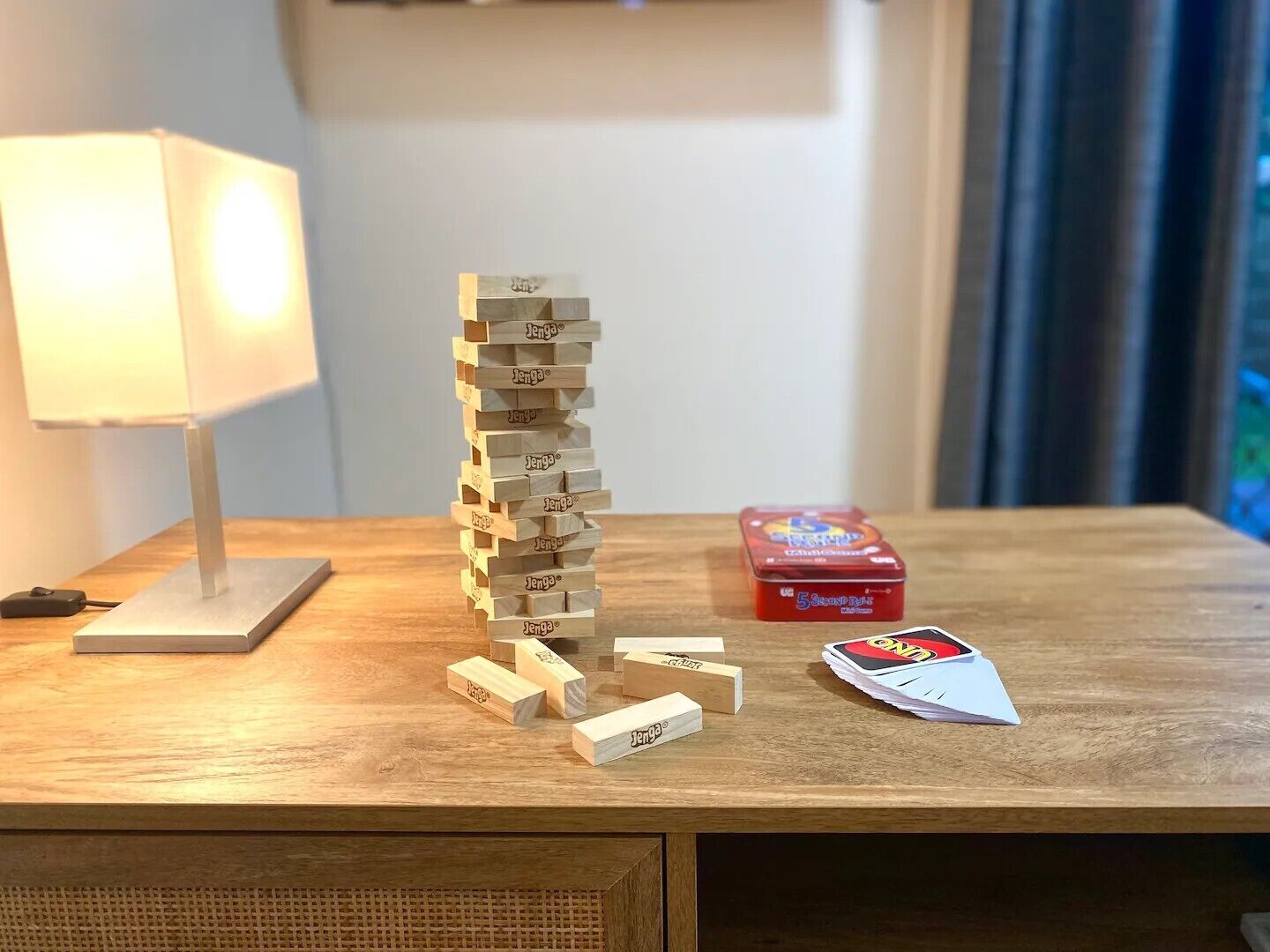 Een houten Jenga-toren op een houten tafel met een lamp en een doos Jenga-kaarten.