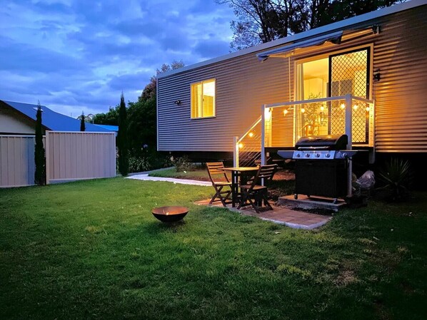 A backyard with a grill, a table, and chairs, and a house with a window and string lights.