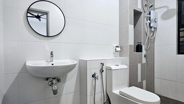 Ein modernes Badezimmer mit Waschbecken, Toilette und Dusche in Weiß. Ein runder Spiegel befindet sich an der Wand, und ein blauer Mülleimer ist zu sehen.