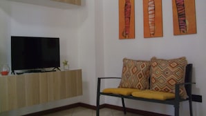 Una sala de estar con un televisor sobre un mueble de madera, un sofá negro con cojines estampados y tres cuadros colgados en la pared.