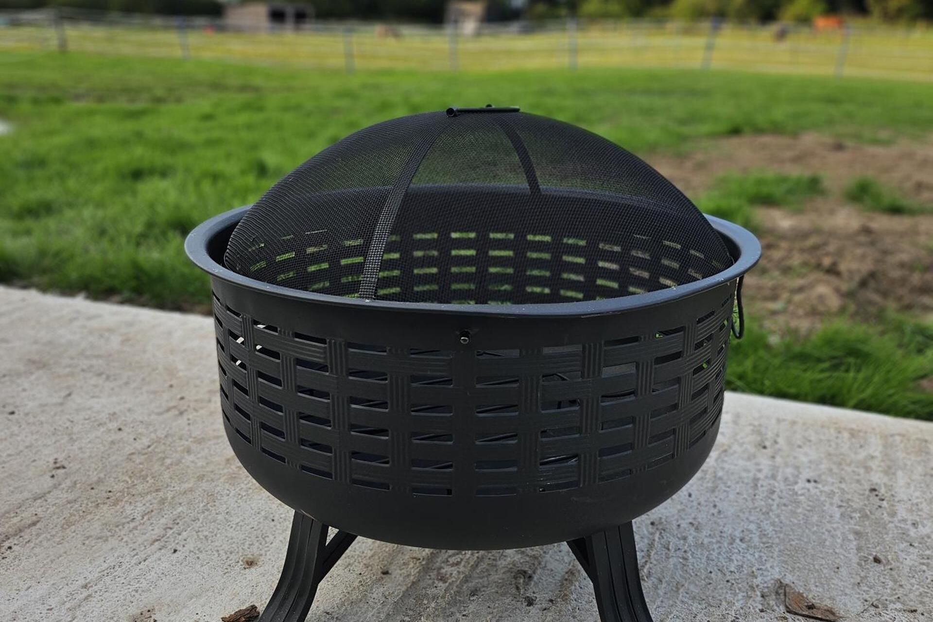 A black outdoor grill with a mesh top, standing on a concrete surface with a grassy field in the background.