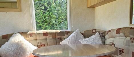 A cozy RV interior with a round table, cushioned seating, and a window with a view of greenery.