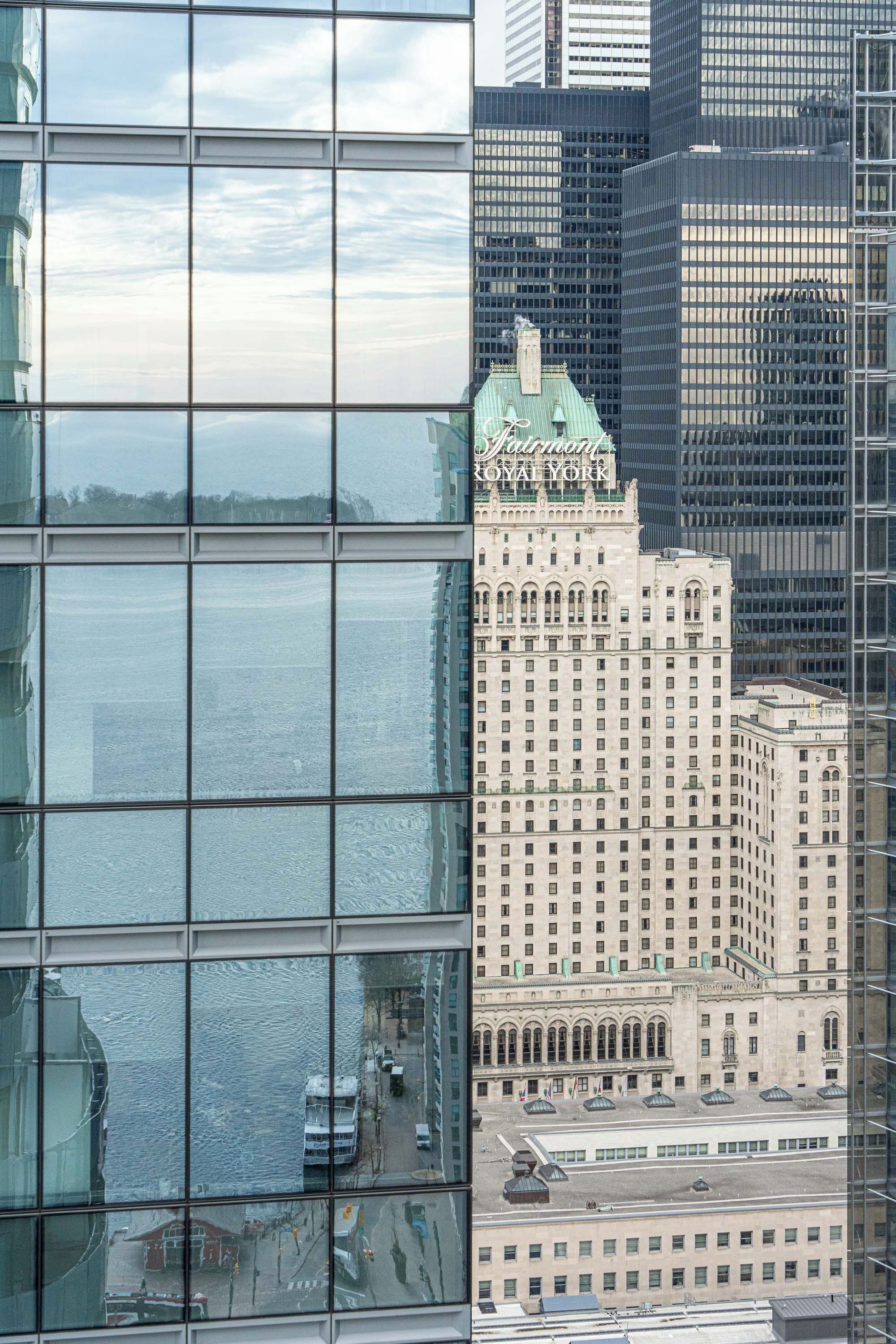 Un edificio de gran altura con fachada de vidrio que refleja el cielo y, al fondo, un edificio histórico con el letrero "Hotel Royal York".