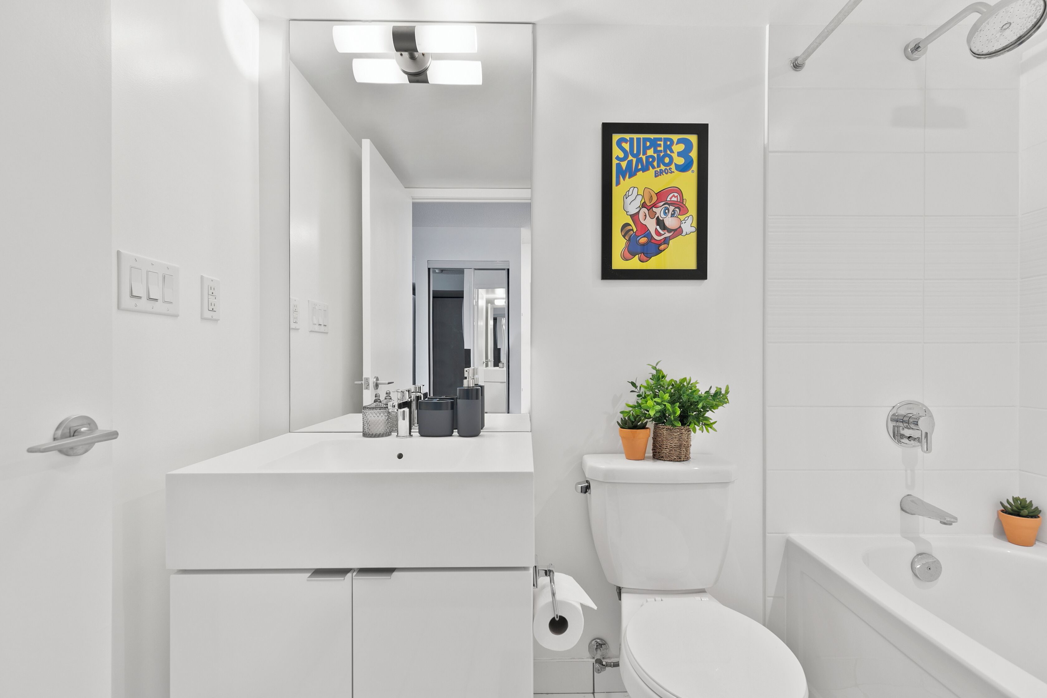 Un baño moderno con lavamanos, inodoro y bañera blancos. Hay un espejo, un dispensador de papel higiénico y un cartel enmarcado de Super Mario Bros. en la pared.