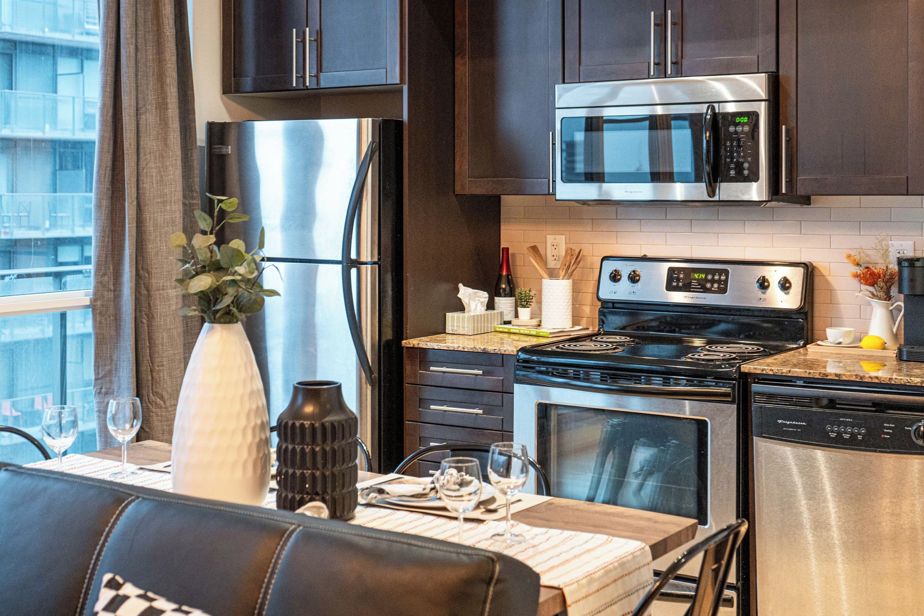 Una cocina moderna con electrodomésticos de acero inoxidable, un comedor con una mesa preparada para dos personas y vistas de la ciudad a través de amplias ventanas.