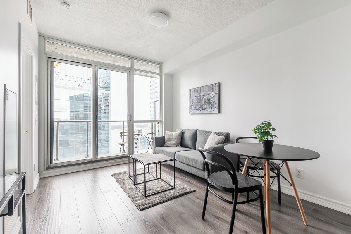 A modern living room with a sofa, coffee table, and chairs, large windows with a city view, and a small table with a plant.