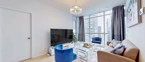 A modern living room with a flat-screen TV, a blue chair, a brown sofa, and large windows with curtains.