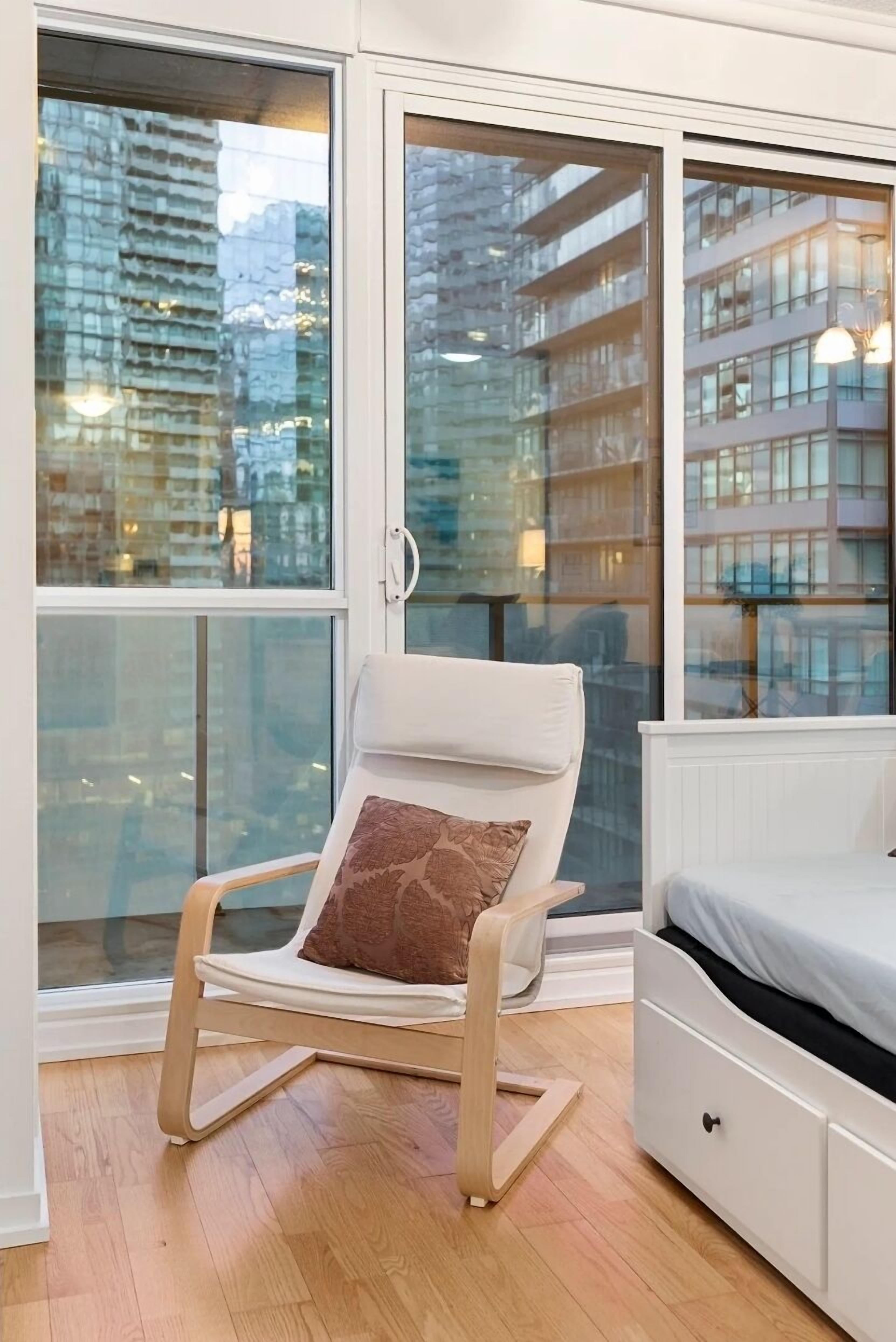 Un dormitorio con una cama blanca, una mecedora de madera con un cojín marrón y vistas a edificios de la ciudad a través de amplios ventanales.