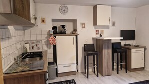 Cocina compacta con fregadero, estufa y cafetera. Hay dos taburetes de bar y un televisor pequeño sobre un pedestal.