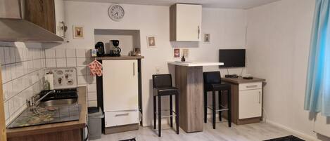 Cocina compacta con fregadero, estufa y cafetera. Hay dos taburetes de bar y un televisor pequeño sobre un pedestal.