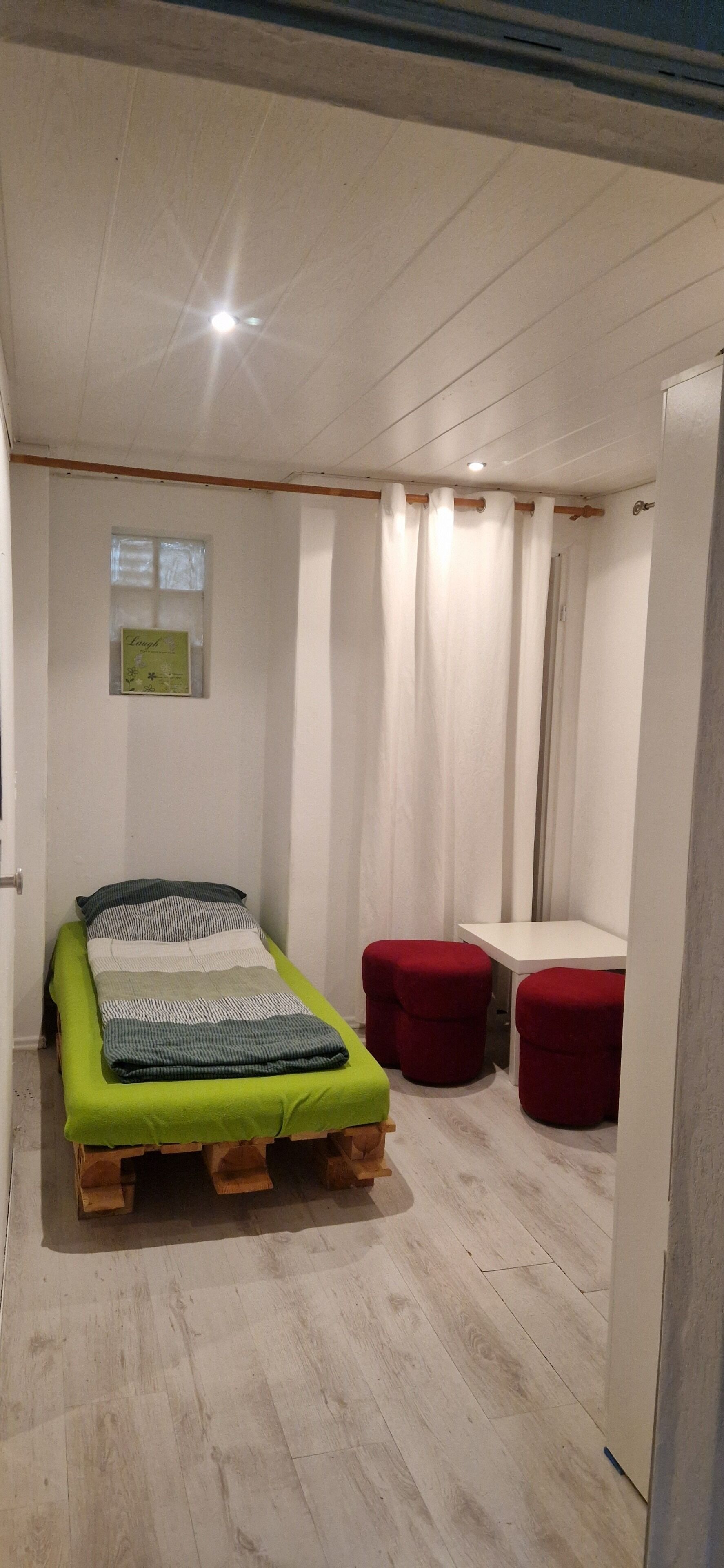 Ein kleines Zimmer mit einem Einzelbett, einem kleinen Tisch und zwei roten Hocker.