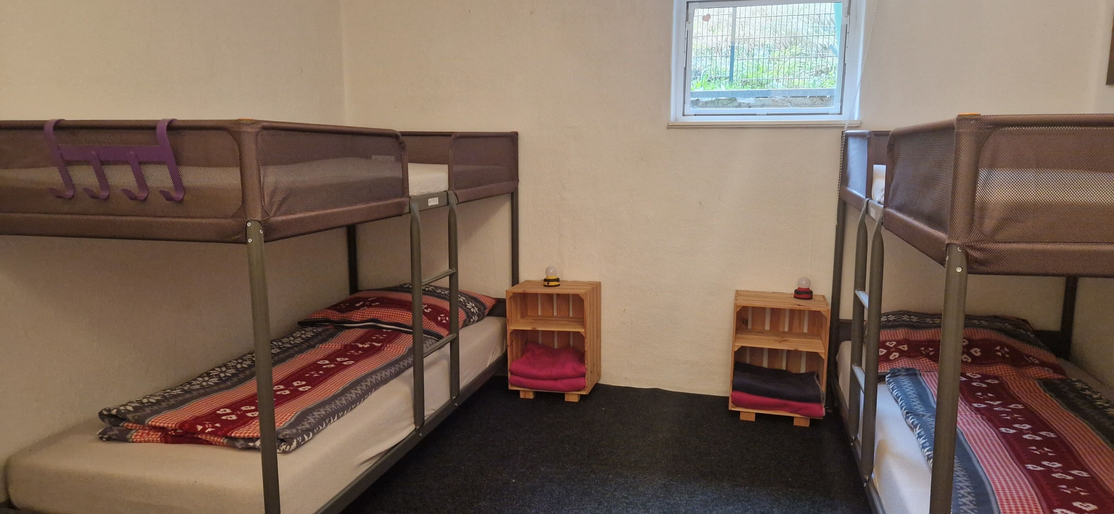 A dormitory room with bunk beds, two wooden nightstands, and a window with a view of greenery.