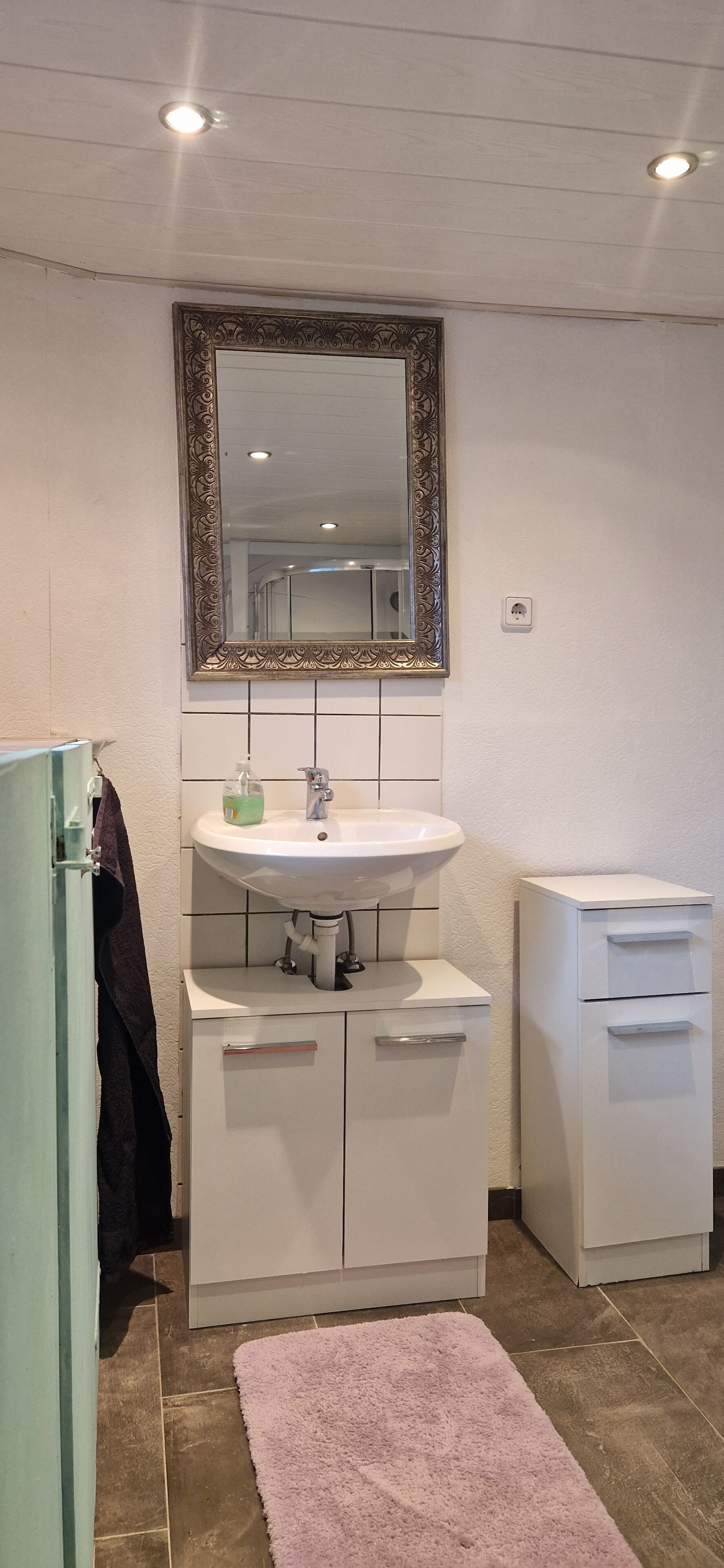 Ein Badezimmer mit einem weißen Waschbecken, einem Spiegel mit aufwendigem Rahmen, einem rosa Teppich und einem Schrank mit Schubladen.