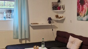 Una habitación acogedora con sofá y dos camas individuales hechas de cajas de madera, una mesita con una botella, una lámpara y una estantería con objetos decorativos.