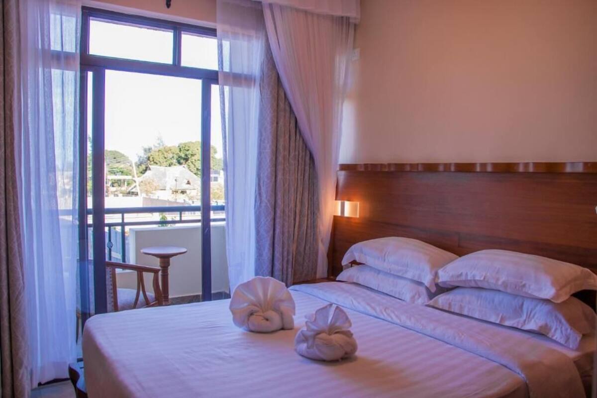 A hotel room with a bed, pillows, a bedside table, and a view through a large window.
