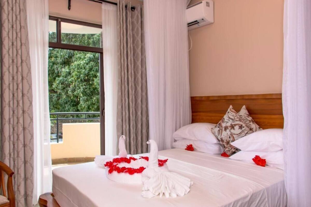 A hotel room with a bed, white curtains, a wooden headboard, and a view of greenery outside.