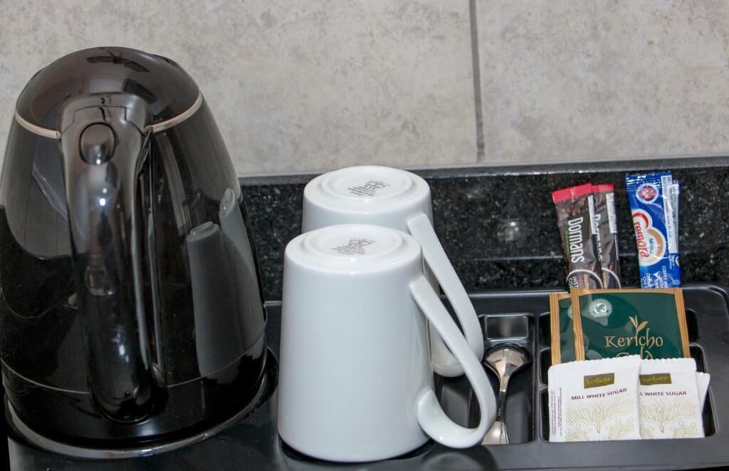 Uma estação de café com uma cafeteira de café preto, duas canecas brancas, uma colher, um sachê de açúcar e um sachê de leite.