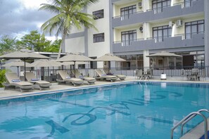 A swimming pool with lounge chairs and umbrellas, surrounded by a building with multiple balconies and air conditioning units. - Hotel Comster Mtwapa (Mtwapa)